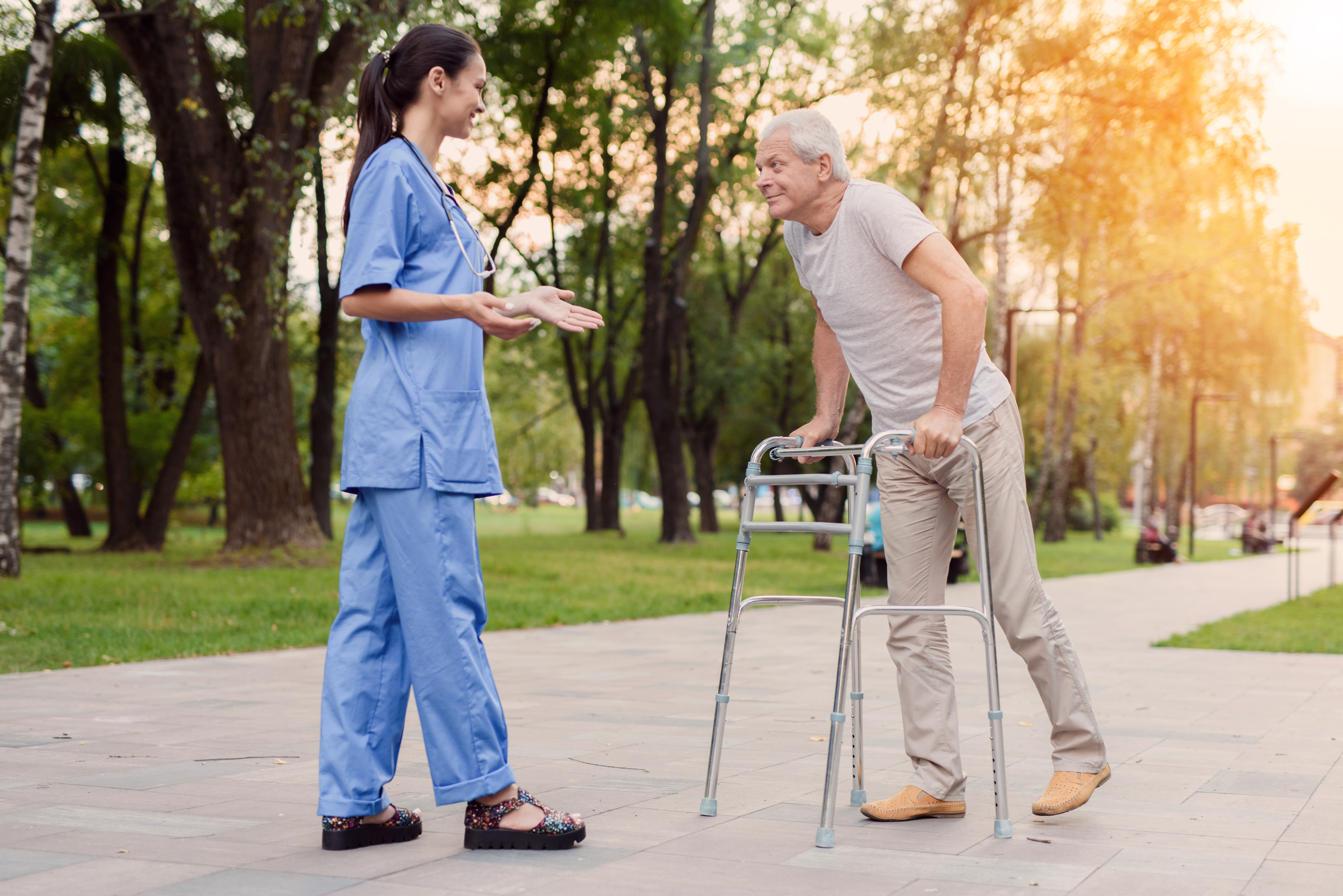 A young nurse is standing in the park, with an elderly man and is helping him walk.